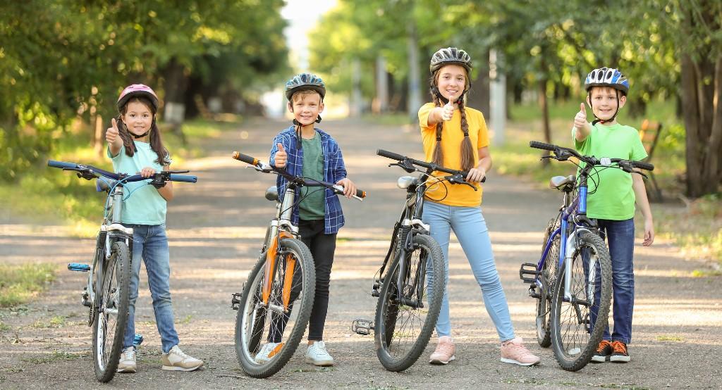 Bukósisakos, mosolygós gyerekek biciklivel állnak egy szép, fákkal szegélyezett úton likeot mutatva ujjakkal 