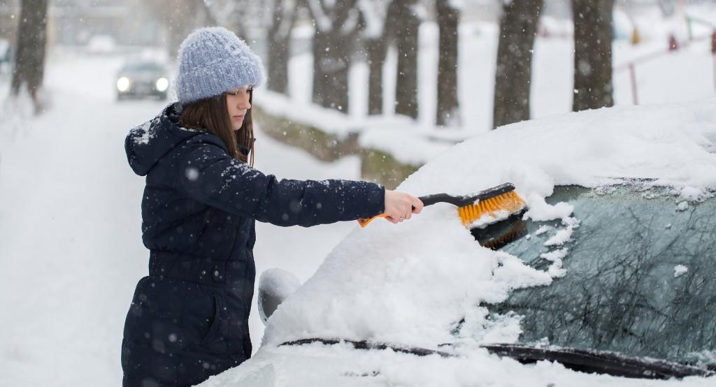 Havas környezetben az autó mellett álló nő kézi seprűvel hó mentesíti a kocsi szélvédőjét
