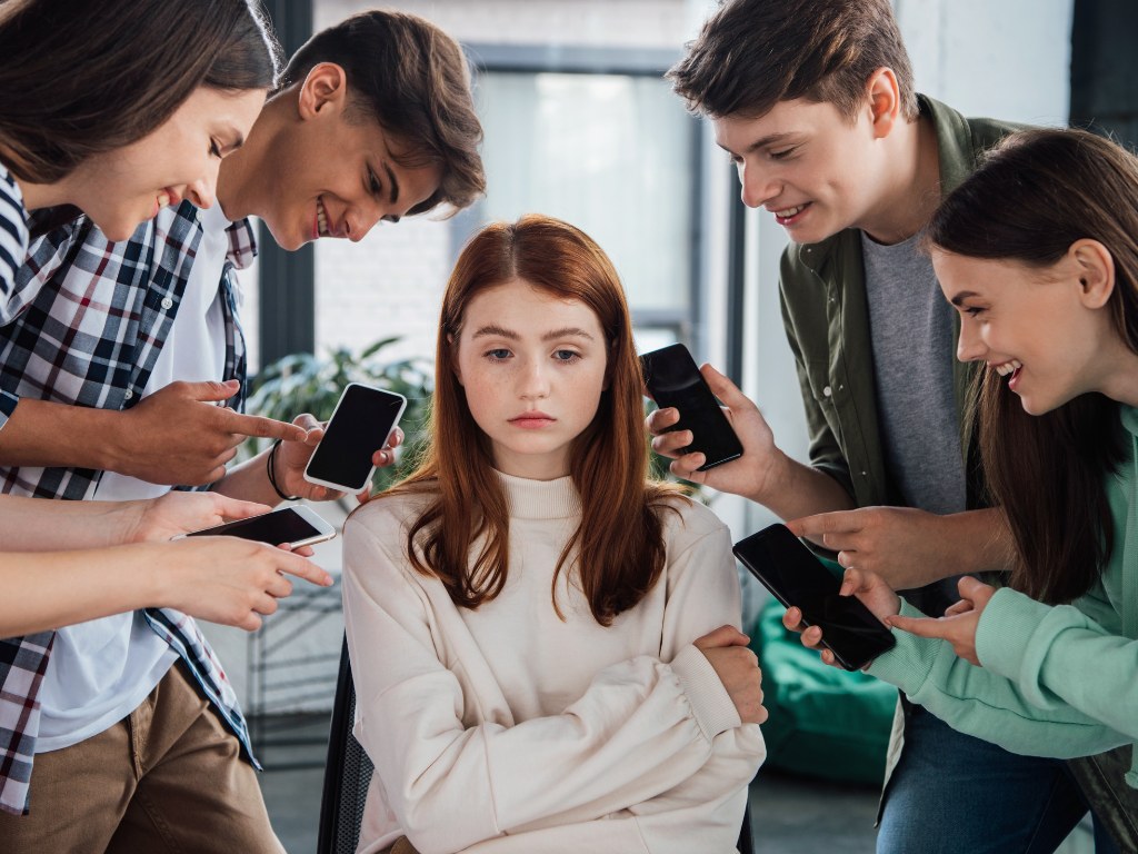 Széken ülő szomorú lányt vesz körül több kamasz a telefonjukat mutogatva neki