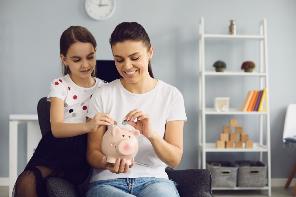 Anya és lánya ül a fotelben és aprót dob egy malac perselybe.