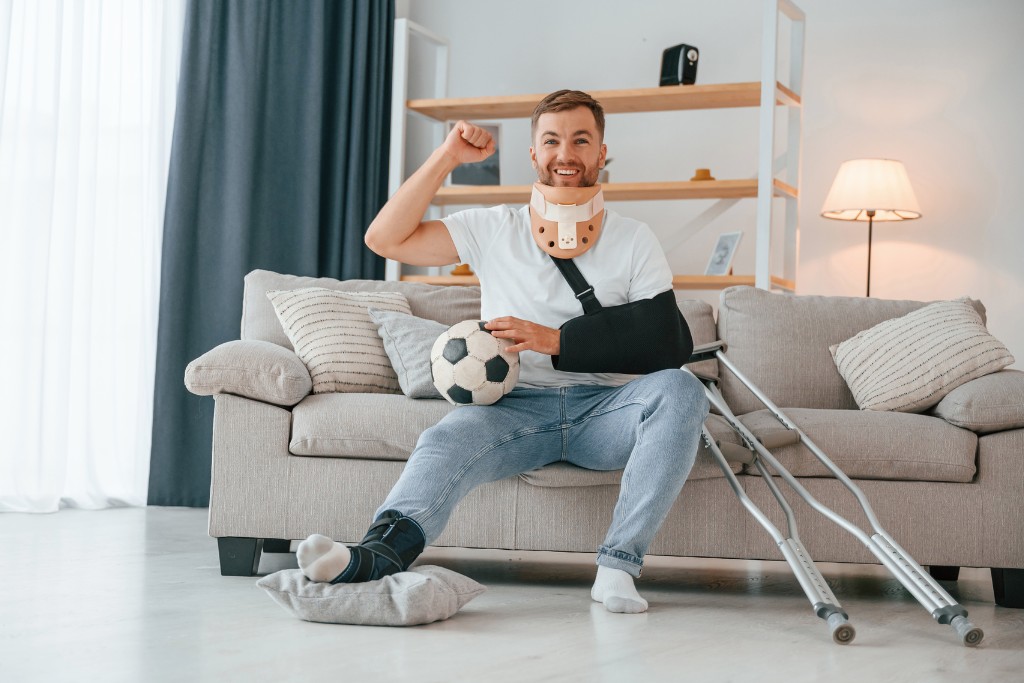 Férfi mosolyogva ül a kanapén. Karja, nyaka, lába gipszben, mellette makók.